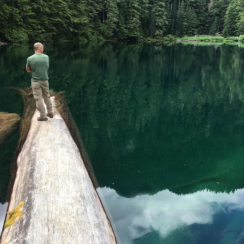 Craig Holt from behind standing on a log in the middle of a lake surrounded by evergreen forests